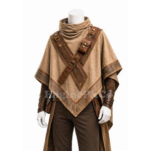 May include: A tan poncho with brown leather accents, straps, and a small pouch. The poncho has a high collar and is paired with brown pants and arm cuffs. The outfit is displayed on a mannequin.