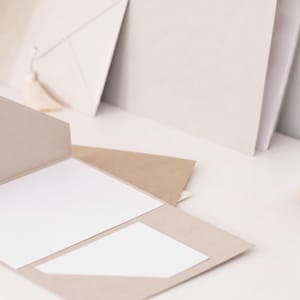 May include: A light brown card with a white insert is open, with a stack of similar cards and envelopes in the background. A wooden stamp with a black handle is on the right. The stationery is on a white surface.