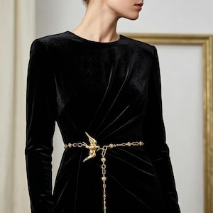 Puede incluir: Un vestido de terciopelo negro con mangas largas, ceñido a la cintura con un cinturón de cadena dorada. El cinturón presenta un colgante en forma de pájaro y una cadena colgante con cuentas decorativas. El vestido tiene un diseño clásico y elegante.