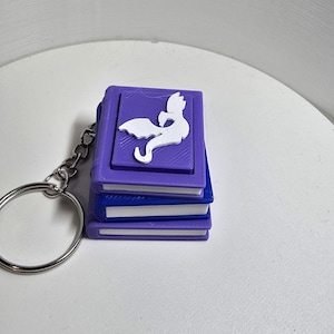 May include: A keychain featuring a stack of three miniature books with purple and blue covers and white pages. The top book cover displays a raised white silhouette of a dragon with wings. A silver metal ring and chain are attached to the side.