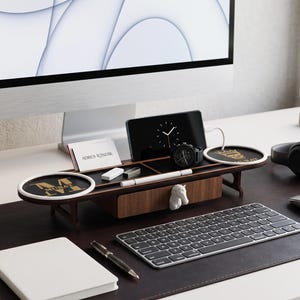 May include: A dark wood desk organizer featuring a central drawer with a white horse head handle, two circular wireless charging pads with gold logos, a smartphone stand, a business card slot, and a pen tray, positioned on a dark leather desk mat.