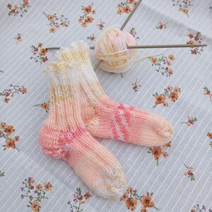 Peut inclure: Une paire de chaussettes tricotées à la main, aux tons pêche, rose et blanc. Elles reposent sur un tissu à motifs floraux. On aperçoit une pelote de laine et des aiguilles à tricoter. La laine est un mélange de jaune, rose et blanc.