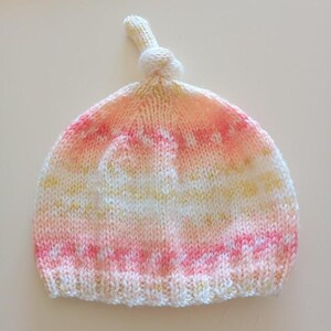 Peut inclure: Un bonnet de bébé tricoté à la main aux couleurs pastel. Le chapeau présente un dessus noué et un mélange de fil rose, jaune et blanc. Le bonnet est conçu avec un bord côtelé et une couronne arrondie, idéal pour un nouveau-né.