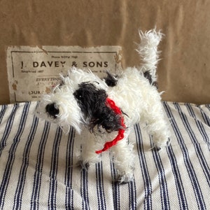 May include: A white and black fluffy toy dog with a red collar. The dog is standing on a striped fabric surface.