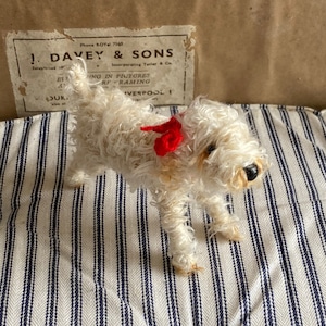 May include: A white, fluffy toy dog with a red bow around its neck. The dog is sitting on a striped fabric surface.