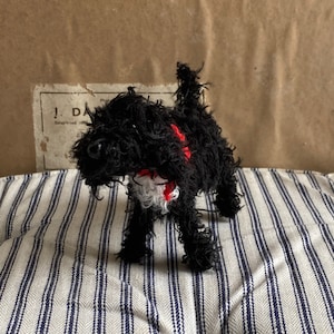 May include: Black plush toy dog with a red and white collar. The dog is standing on a striped fabric surface.