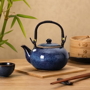 Pode incluir: Uma chaleira de cerâmica azul com alça e tampa pretas, sobre uma esteira de bambu. A chaleira tem um acabamento brilhante e um bico. Uma xícara pequena é visível à esquerda.
