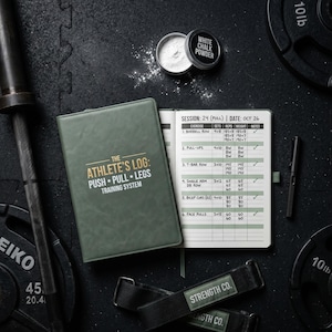 May include: An open training log book with a green cover and the text "THE ATHLETE'S LOG: PUSH • PULL • LEGS TRAINING SYSTEM" sits next to a pen and a container of white chalk powder. Weightlifting straps and a barbell are also visible.