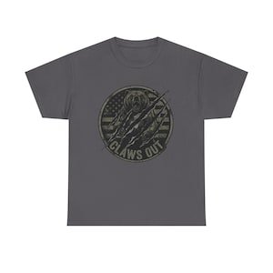 Pode incluir: Uma t-shirt de manga curta cinzento-carvão com um gráfico circular no peito. O design apresenta um urso com marcas de garras sobre um padrão de bandeira americana desgastada, com o texto CLAWS OUT impresso em negrito na borda inferior.