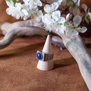 May include: A silver ring with a square face and a large, round, blue stone. The ring is displayed on a small, light-colored cone-shaped stand. White flowers and a piece of driftwood are in the background.
