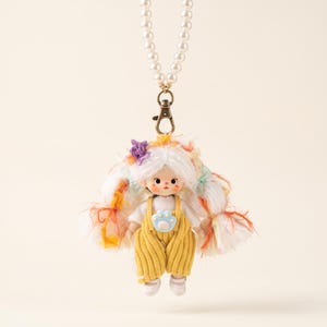 May include: A small doll charm hanging from a white pearl bead chain with a gold-tone lobster clasp. The doll has white hair with orange and yellow accents, wearing yellow knit overalls with a light blue paw print emblem on the chest.