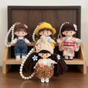 May include: Four small doll keychains with brown hair and distinct outfits. One wears a cream crochet hat, white top, and brown curly skirt. Others wear denim overalls, a yellow knit hat, or a floral kimono, all featuring white pearl bead chains.