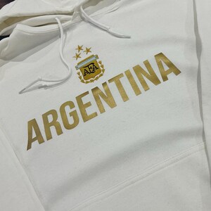 Puede incluir: Sudadera con capucha blanca con la palabra "ARGENTINA" en letras doradas. La sudadera con capucha presenta un emblema dorado con tres estrellas y las letras "AFA". La sudadera con capucha tiene una capucha con cordón y un bolsillo delantero.
