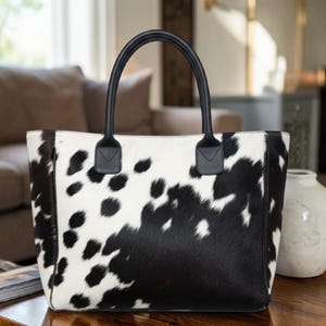 May include: A tote bag with a cowhide pattern in black and white. The bag has black leather handles and trim. The bag is rectangular and appears to be sitting on a wooden surface.