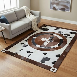 May include: Rectangular area rug with a cowhide pattern and a central circular design. The rug features a brown border and a central emblem of a bird. The rug is placed on a hardwood floor.