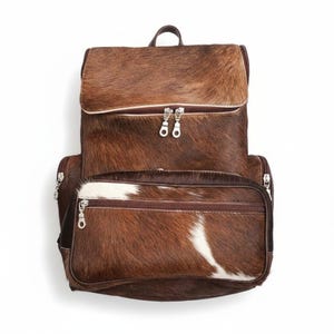 Puede incluir: Mochila de piel de vaca marrón y blanca con cierre de solapa y múltiples compartimentos con cremallera. La bolsa tiene un asa superior y cremalleras plateadas. La mochila está hecha de un material natural con un patrón único.
