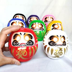 May include: A collection of colorful Daruma dolls, each with a unique color scheme and facial features. The red doll in the foreground has gold calligraphy. The dolls are round and have a traditional Japanese design.