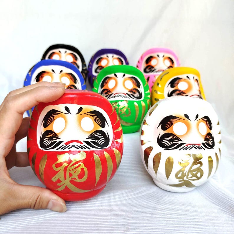 May include: A collection of colorful Daruma dolls, each with a unique color scheme and facial features. The red doll in the foreground has gold calligraphy. The dolls are round and have a traditional Japanese design.