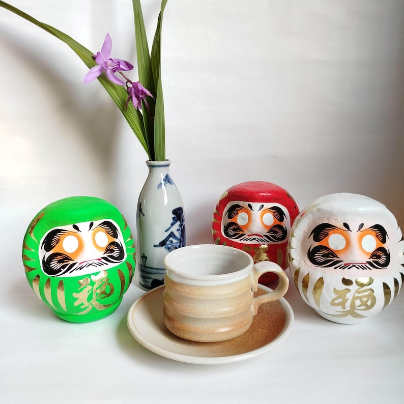 May include: A still life featuring Japanese Daruma dolls in green, red, and white, alongside a ceramic cup and saucer. A small vase holds a sprig of purple flowers and green leaves. The Daruma dolls have gold accents.