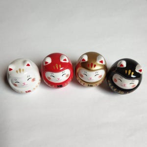 以下が含まれることがあります： 4つの日本のダルマ人形が並んでいます。人形は卵形で猫の顔をしており、白、赤、金、黒で塗装されています。各人形は笑顔で、下部には「幸運」の漢字が書かれています。