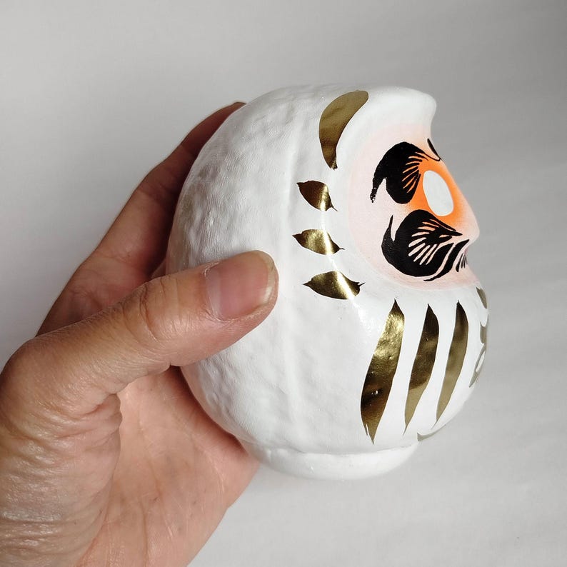 May include: White Daruma doll with gold accents. The doll features a painted face with orange, black, and white details. Gold leaf-like shapes adorn the body. The Daruma doll is a traditional Japanese good luck charm.