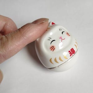 以下が含まれることがあります： 白いセラミック製の招き猫の置物。笑顔の顔、黒い目、赤いアクセントが特徴です。日本の幸運の猫は金色の装飾と赤い「福」の文字が施されています。この置物は幸運の象徴です。
