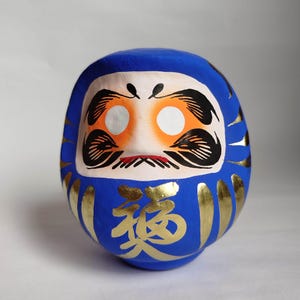 May include: A blue Daruma doll with a white face, orange accents, and black details. Gold Japanese characters are painted on the front. The doll has a round shape and is a traditional Japanese good luck charm.