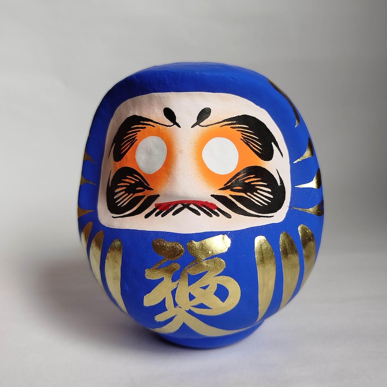 May include: A blue Daruma doll with a white face, orange accents, and black details. Gold Japanese characters are painted on the front. The doll has a round shape and is a traditional Japanese good luck charm.