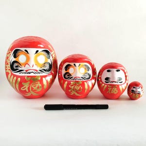 May include: Four red Daruma dolls of different sizes, featuring white, black, and gold details. Each doll has a stylized face and gold Japanese characters. A black marker is next to the dolls for scale.