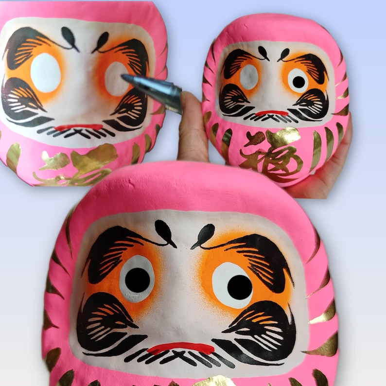 May include: Three pink Daruma dolls with black, gold, and white details. The dolls have large eyes, eyebrows, and a mustache. One doll is being held by a hand with a pen pointing at it. The dolls have gold Japanese characters.