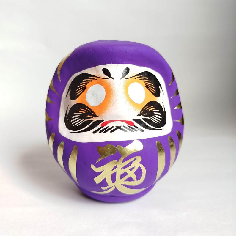 May include: A purple Daruma doll with a white face, orange eye accents, and black details. Gold Japanese characters are on the front. Gold accents are on the sides. This traditional Japanese doll is a symbol of good luck.