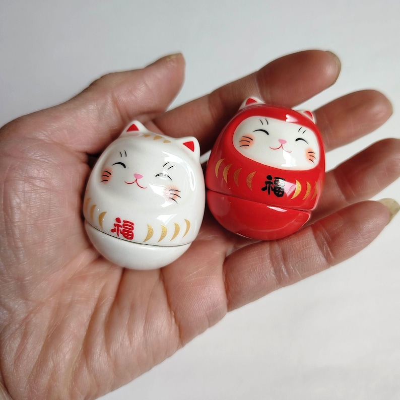 以下が含まれることがあります： 2つの小さな猫の形をした置物。1つは金色のアクセントと「幸運」の漢字が入った白。もう1つは黒い文字が入った赤。どちらも笑顔で、装飾品である可能性が高いです。