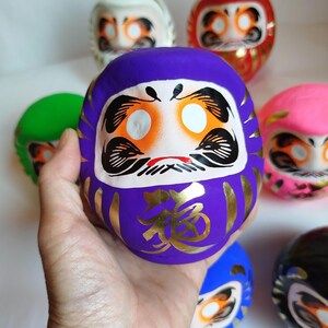 May include: A purple Daruma doll with gold accents and the Japanese character for "fortune." The doll has white eyes, orange and white facial details, and a red mustache. Other colorful Daruma dolls are in the background.