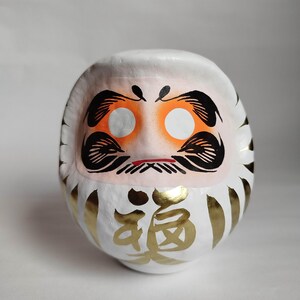 May include: A white Daruma doll with orange eye sockets, black eyebrows and mustache, and a red mouth. The doll has gold accents and Japanese characters. This traditional Japanese doll is a symbol of good luck and perseverance.