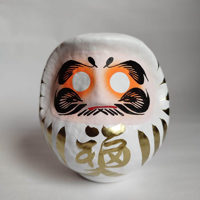 May include: A white Daruma doll with orange eye sockets, black eyebrows and mustache, and a red mouth. The doll has gold accents and Japanese characters. This traditional Japanese doll is a symbol of good luck and perseverance.