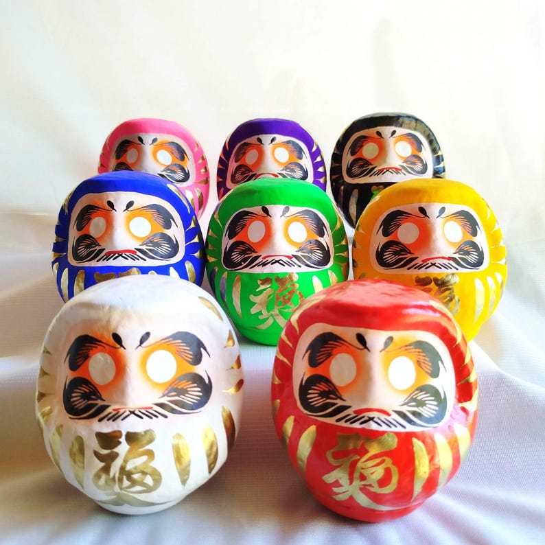 May include: A collection of Daruma dolls in various colors, including red, yellow, green, blue, purple, pink, black, and white. Each doll features a stylized face with eyes, a mustache, and gold accents. Some dolls have gold Japanese characters.