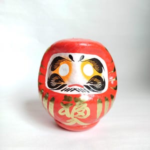 May include: A red Daruma doll with gold accents and Japanese characters. The doll has a white face with black and gold details, including eyes, eyebrows, and a mustache. The Daruma is a traditional Japanese good luck charm.