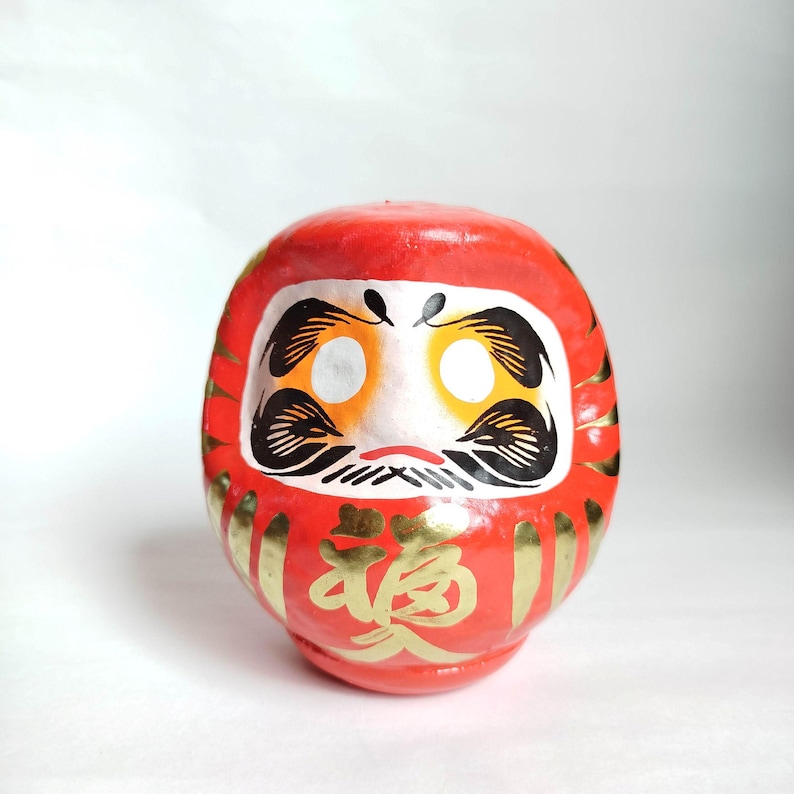 May include: A red Daruma doll with gold accents and Japanese characters. The doll has a white face with black and gold details, including eyes, eyebrows, and a mustache. The Daruma is a traditional Japanese good luck charm.