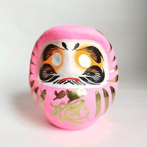 May include: A pink Daruma doll with a white face, black eyebrows, and a red mouth. The doll has gold accents and Japanese characters on the front. The Daruma doll is a traditional Japanese good luck charm.