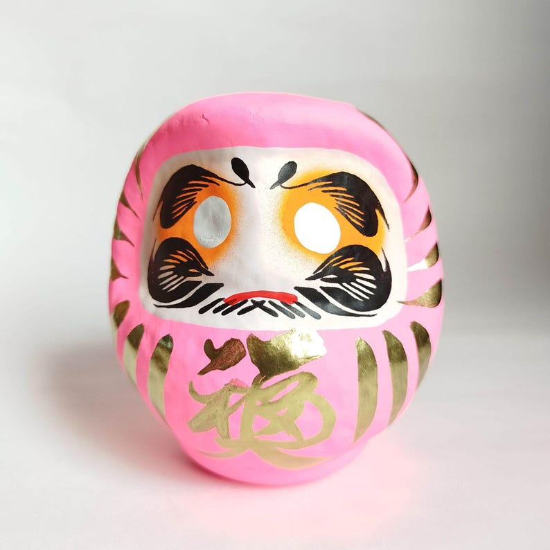 May include: A pink Daruma doll with a white face, black eyebrows, and a red mouth. The doll has gold accents and Japanese characters on the front. The Daruma doll is a traditional Japanese good luck charm.