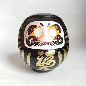 May include: A black Daruma doll with gold accents and Japanese characters. The doll has a white face with orange cheeks, black eyebrows, and a small red mouth. Gold stripes adorn the sides.