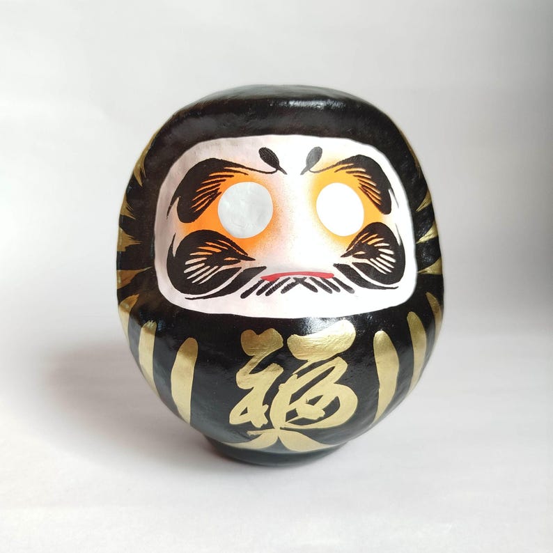 May include: A black Daruma doll with gold accents and Japanese characters. The doll has a white face with orange cheeks, black eyebrows, and a small red mouth. Gold stripes adorn the sides.
