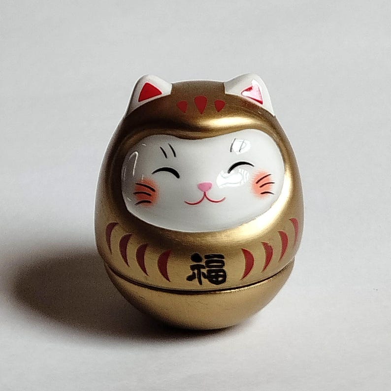 以下が含まれることがあります： 白い顔と赤いアクセントが付いた金色のダルマ人形。人形は笑顔で、黒い目と赤い頬をしています。「幸運」の漢字が前面に印刷されています。