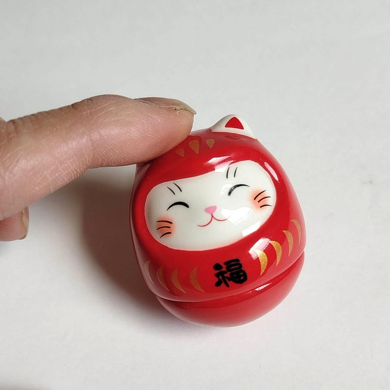 以下が含まれることがあります： 赤いダルマ人形。猫のような顔で、黒い目、ひげ、笑顔の口が特徴です。人形には金のアクセントがあり、前面には「幸運」の漢字が書かれています。