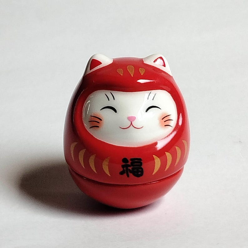 以下が含まれることがあります： 猫のようなデザインの赤いダルマ人形。人形は白い顔で、笑顔、ピンクのひげがあり、下半分には「幸運」を表す日本語の文字があります。人形の耳は白で、金色のアクセントが付いています。