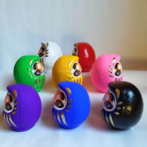 May include: A collection of Daruma dolls in various colors, including green, white, red, yellow, pink, purple, blue, and black. Each doll features a detailed face with gold accents and a rounded shape, representing good luck and perseverance.
