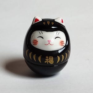 以下が含まれることがあります： 黒いダルマ人形、猫のような顔。人形は白い顔で、笑顔、ピンクの頬、ピンクの鼻。耳は白で赤い先端。前面には金色の日本語の文字があります。
