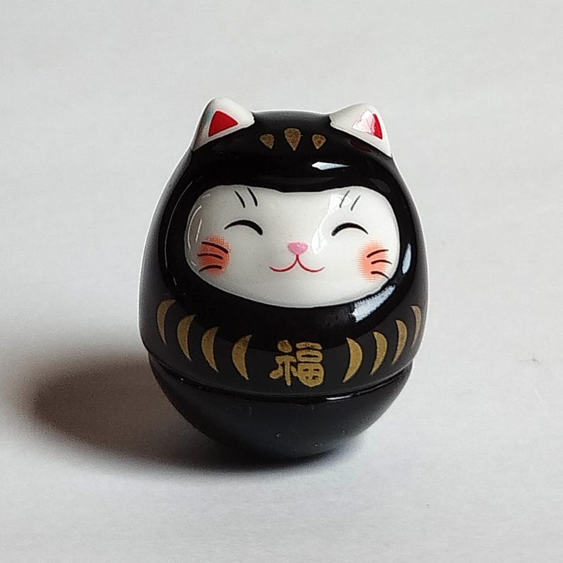以下が含まれることがあります： 黒いダルマ人形、猫のような顔。人形は白い顔で、笑顔、ピンクの頬、ピンクの鼻。耳は白で赤い先端。前面には金色の日本語の文字があります。