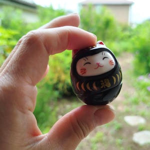 以下が含まれることがあります： 黒と金のダルマ人形、猫の顔のデザイン。人形は赤いアクセントのある白い顔をしており、金色の「幸運」の漢字が描かれています。ぼやけた緑色の背景の前に人形が保持されています。