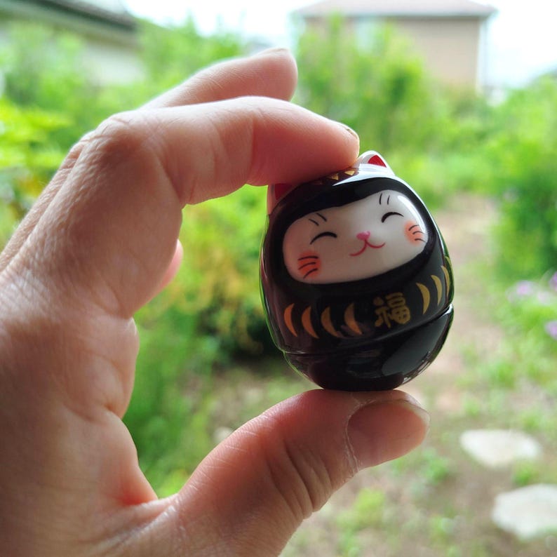 以下が含まれることがあります： 黒と金のダルマ人形、猫の顔のデザイン。人形は赤いアクセントのある白い顔をしており、金色の「幸運」の漢字が描かれています。ぼやけた緑色の背景の前に人形が保持されています。